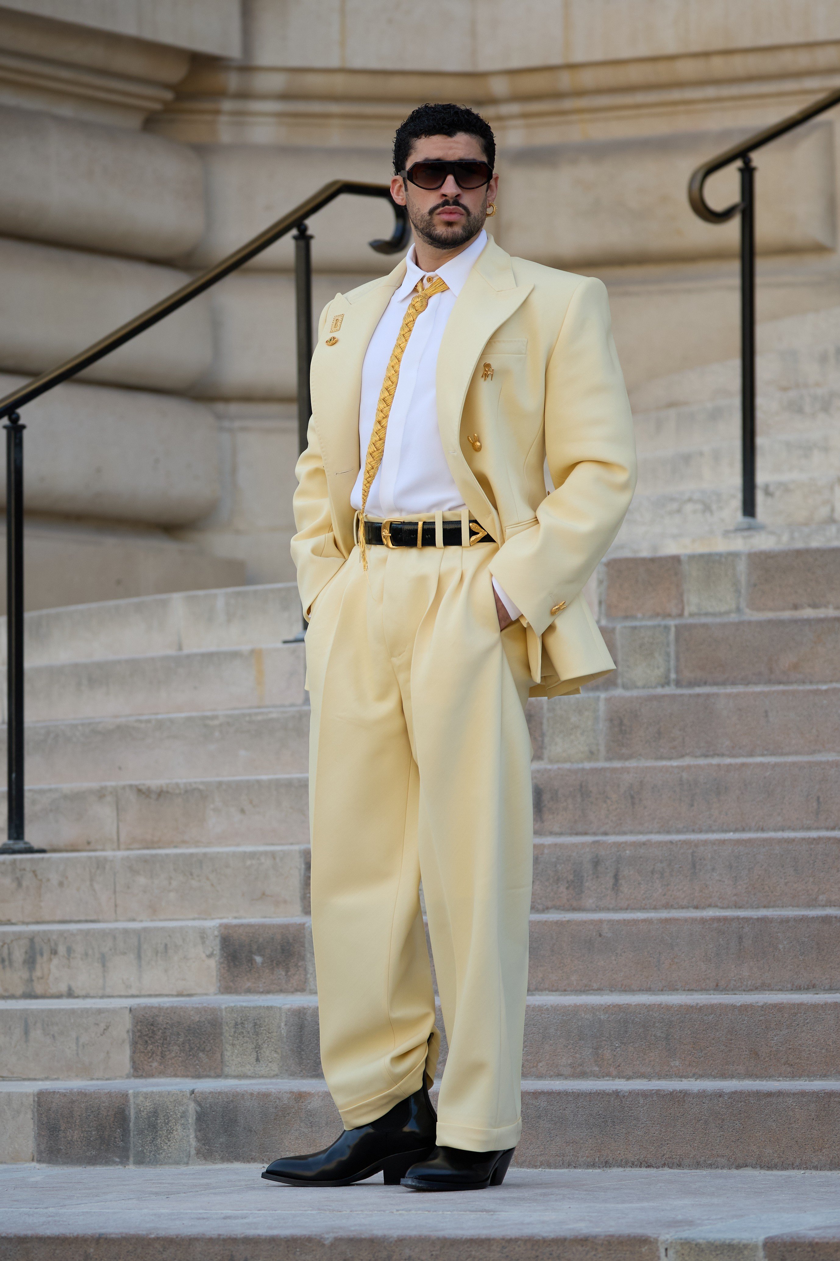 Homem com barba e óculos escuros, usando um terno amarelo-claro, camisa branca, gravata dourada e botas pretas, posa em uma escadaria de pedra com corrimão escuro