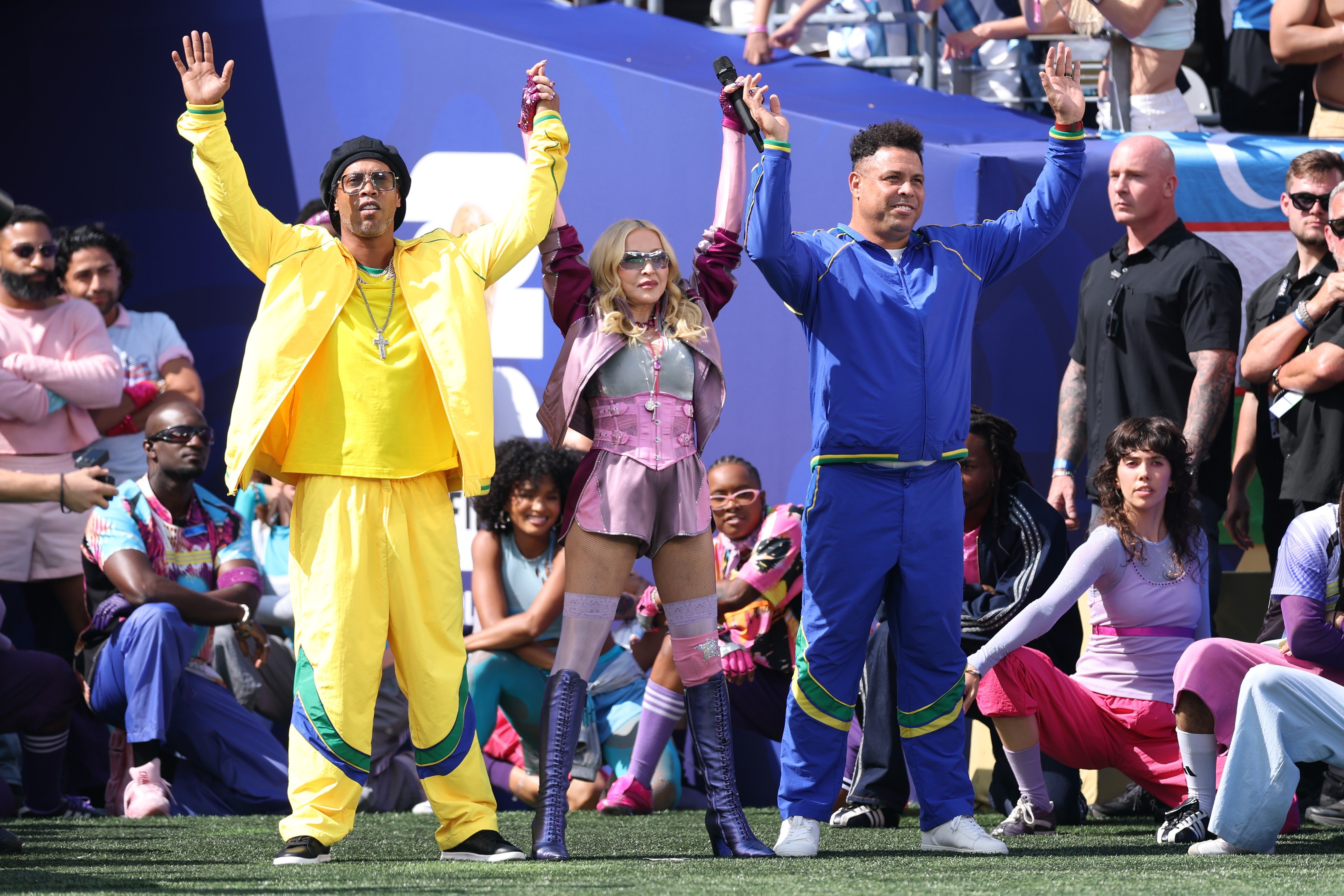 Ronaldinho Gaúcho, Madonna e Ronaldo Fenômeno no palco, braços erguidos, celebrando. Ronaldinho veste agasalho amarelo, Madonna um conjunto rosa com botas, e Ronaldo um agasalho azul. O público e seguranças estão ao fundo