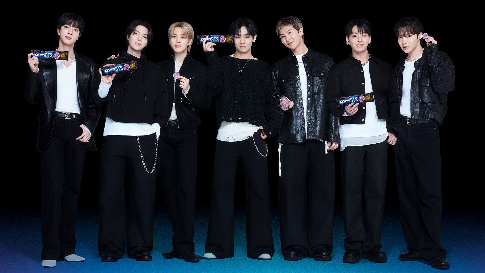 Sete homens jovens, membros do grupo BTS, posam sorrindo em fila, vestindo roupas pretas e brancas. Alguns seguram embalagens de chocolate roxas e pretas com o logo BTS, outros seguram pequenos objetos redondos roxos. O fundo é preto com uma faixa azul na parte inferior
