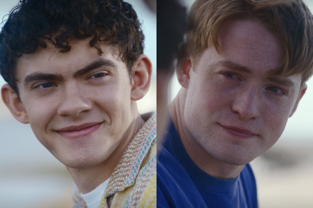 Dois jovens sorrindo, o da esquerda com cabelo cacheado escuro e camisa estampada, o da direita ruivo com sardas e camiseta azul