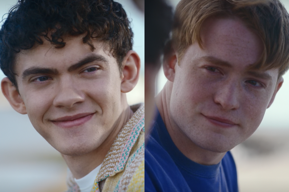 Dois jovens sorrindo, o da esquerda com cabelo cacheado escuro e camisa estampada, o da direita ruivo com sardas e camiseta azul
