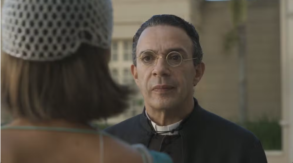 Homem de meia-idade, com óculos redondos e cabelo escuro penteado para trás, veste batina preta e colarinho clerical branco, olhando para uma mulher de costas com chapéu de crochê claro