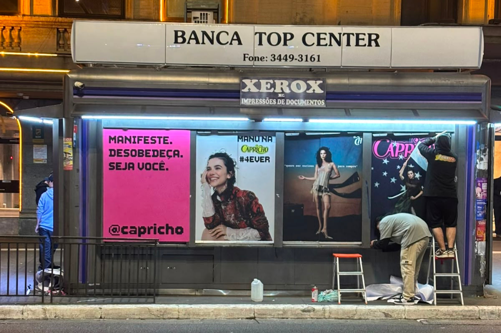 Dois homens instalam pôsteres da revista Capricho em um painel de rua à noite. Um homem de moletom preto está em uma escada, e outro de camisa cinza se curva sobre um rolo de papel. Os pôsteres exibem mulheres jovens, um deles com a frase Manifeste. Desobedeça. Seja você