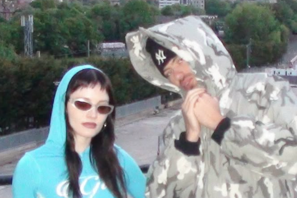 Uma mulher de capuz azul e óculos escuros ao lado de um homem com capuz camuflado e boné preto, em um telhado urbano