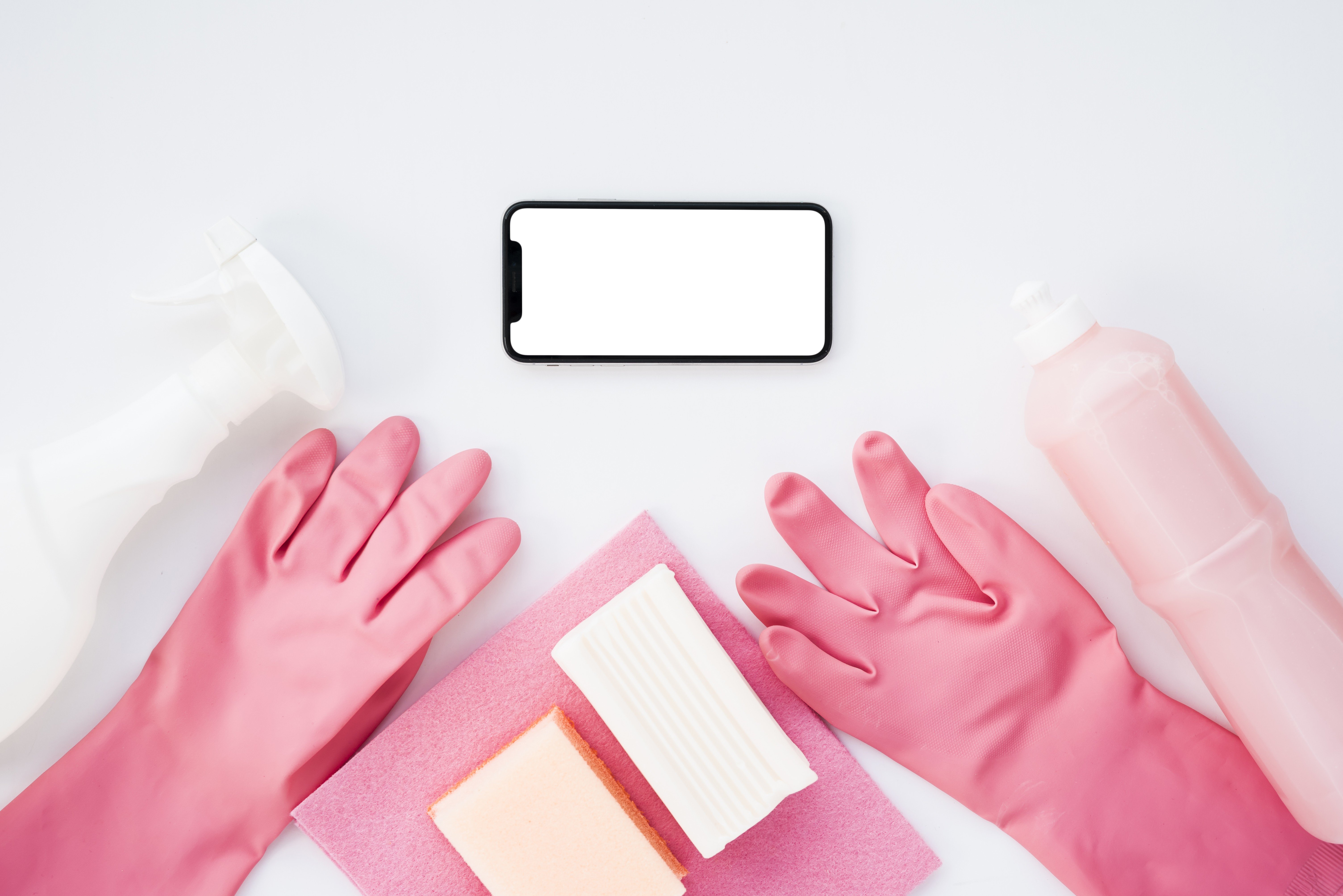 Um smartphone preto com tela branca vazia centralizado, ladeado por luvas de borracha rosa, produtos de limpeza e esponjas sobre fundo branco. À esquerda, um borrifador branco e uma luva rosa. Abaixo do celular, um pano rosa, uma esponja amarela e uma esponja branca. À direita, uma garrafa rosa de produto de limpeza e outra luva rosa