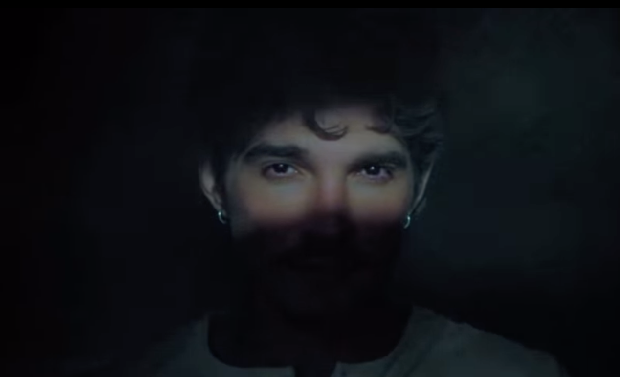Rosto de um homem jovem com cabelo escuro e encaracolado, olhando fixamente para frente. Seus olhos são iluminados, enquanto o restante do rosto está na sombra, criando um efeito dramático. Ele usa brincos de argola prateados