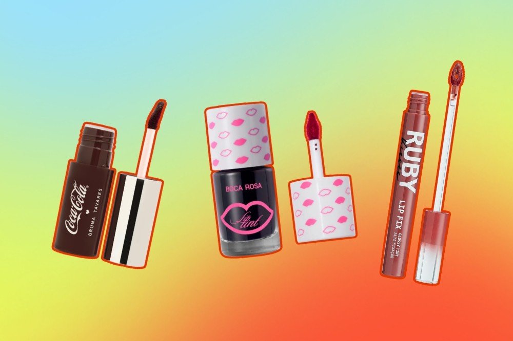Três batons líquidos de marcas diferentes, com seus aplicadores expostos, sobre um fundo degradê de azul, verde e laranja. Da esquerda para a direita: um batom marrom da Bruna Tavares Coca-Cola, um lip tint preto da Boca Rosa e um batom vermelho-tijolo da Ruby Rose Lip Fix