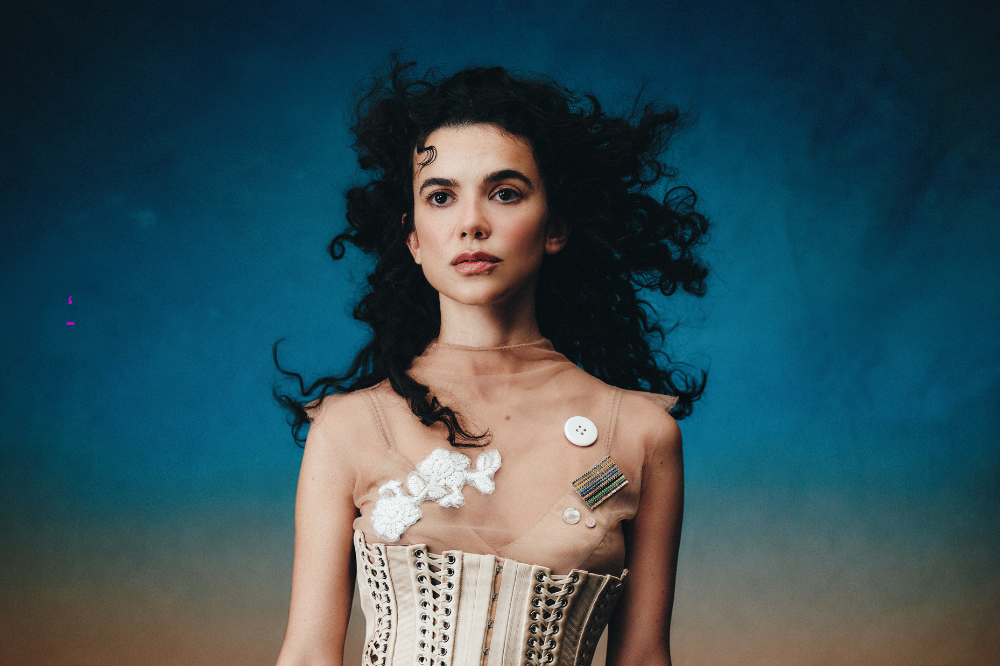 Mulher de cabelos cacheados escuros, com expressão séria, vestindo um espartilho bege e uma blusa transparente com botões e broches, em fundo azul e marrom