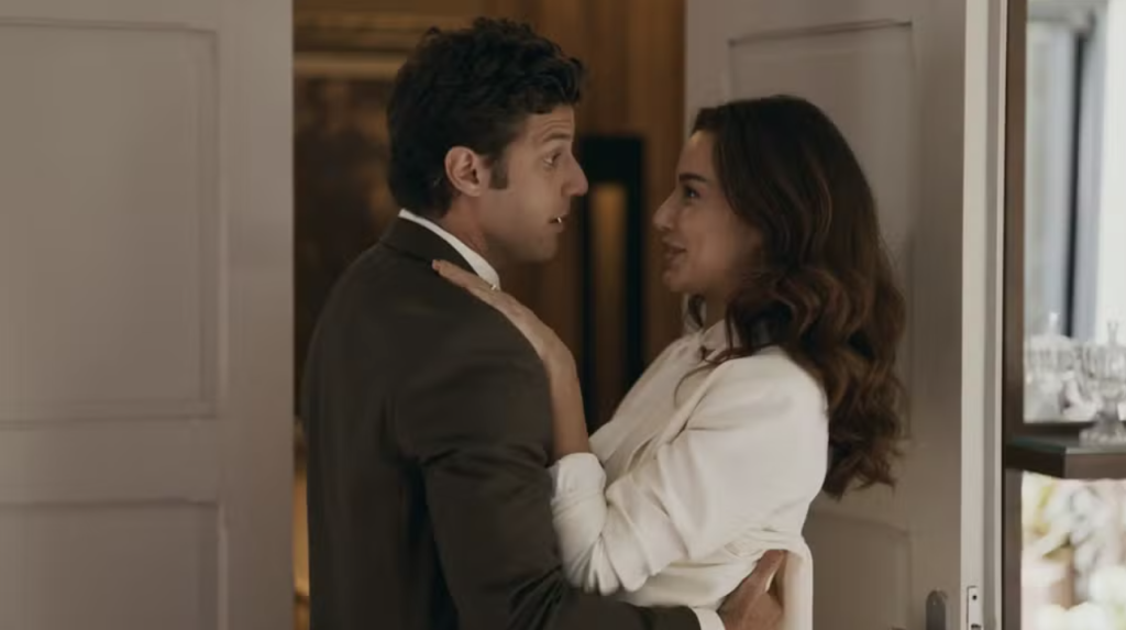 Um homem de terno escuro e uma mulher de blusa branca se abraçam, olhando um para o outro com sorrisos sutis, em um ambiente interno com portas e um espelho ao fundo