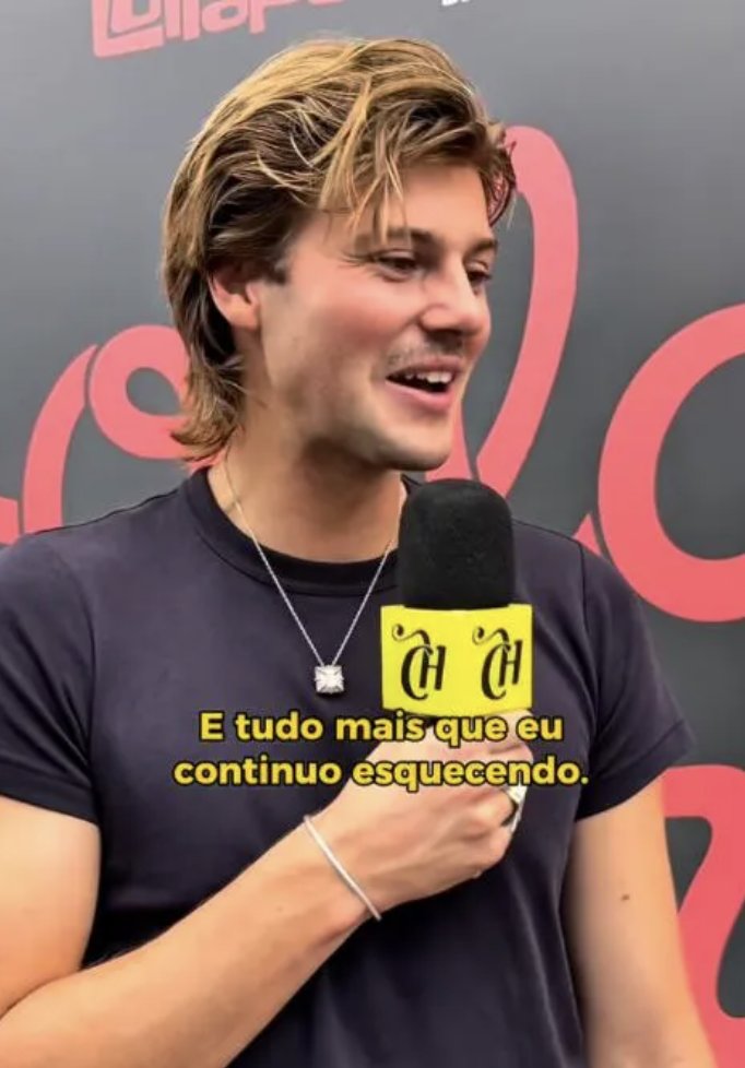 Homem branco de cabelo loiro e bigode, sorrindo, segurando um microfone amarelo com a mão direita. Ele veste uma camiseta azul-marinho e um colar prateado com pingente quadrado. O texto amarelo na imagem diz: E tudo mais que eu continuo esquecendo