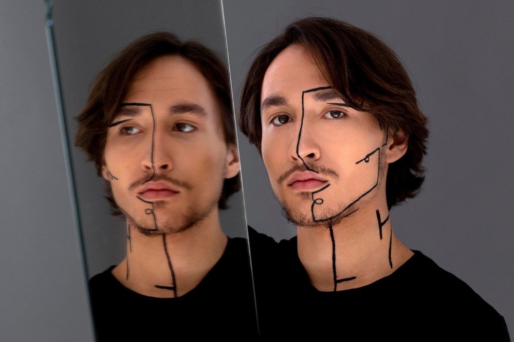 Homem jovem com cabelo castanho e barba rala, usando maquiagem de linhas pretas no rosto e pescoço, olhando para frente com expressão séria, refletido em um espelho à esquerda, sobre fundo cinza