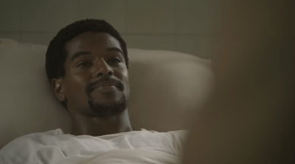 Homem negro deitado, com barba e cabelo crespo, usando camiseta branca, olhando para a direita com um leve sorriso. O fundo é escuro e desfocado