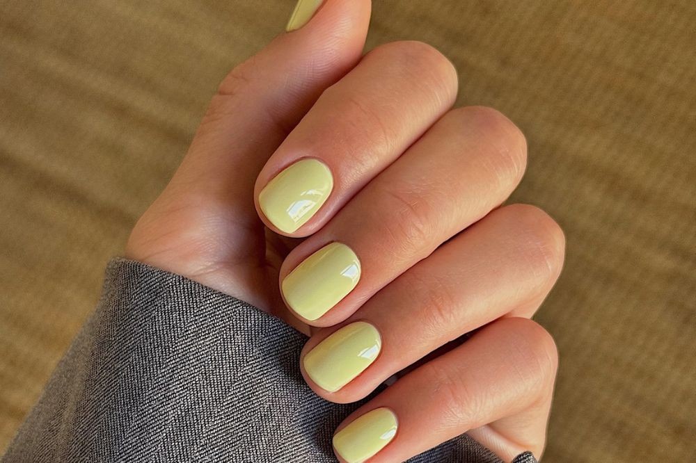 Mão feminina com unhas curtas pintadas de esmalte amarelo pastel brilhante, com a manga de um casaco cinza de tecido texturizado visível no pulso, sobre um fundo marrom claro