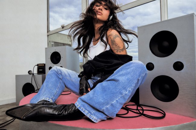 Mulher jovem com cabelos escuros esvoaçantes, usando fones de ouvido, camiseta branca, jaqueta preta, jeans e botas, sentada em uma plataforma rosa entre caixas de som cinzas, com um microfone e amplificador Marshall ao fundo, sob um céu nublado