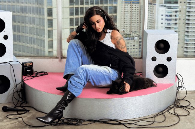 Mulher de cabelos escuros, com tatuagens nos braços, sentada em uma plataforma rosa e cinza, usando fones de ouvido, calça jeans larga, blusa branca, jaqueta preta e botas de cano alto. Ela acaricia um gato preto deitado ao seu lado. Ao redor, há caixas de som prateadas, um microfone e fios espalhados. Ao fundo, uma janela com prédios altos da cidade