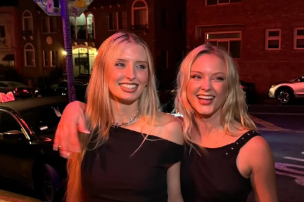 Duas mulheres loiras sorrindo, uma com a mão no ombro da outra, vestindo blusas pretas, em uma rua iluminada à noite com carros e edifícios ao fundo