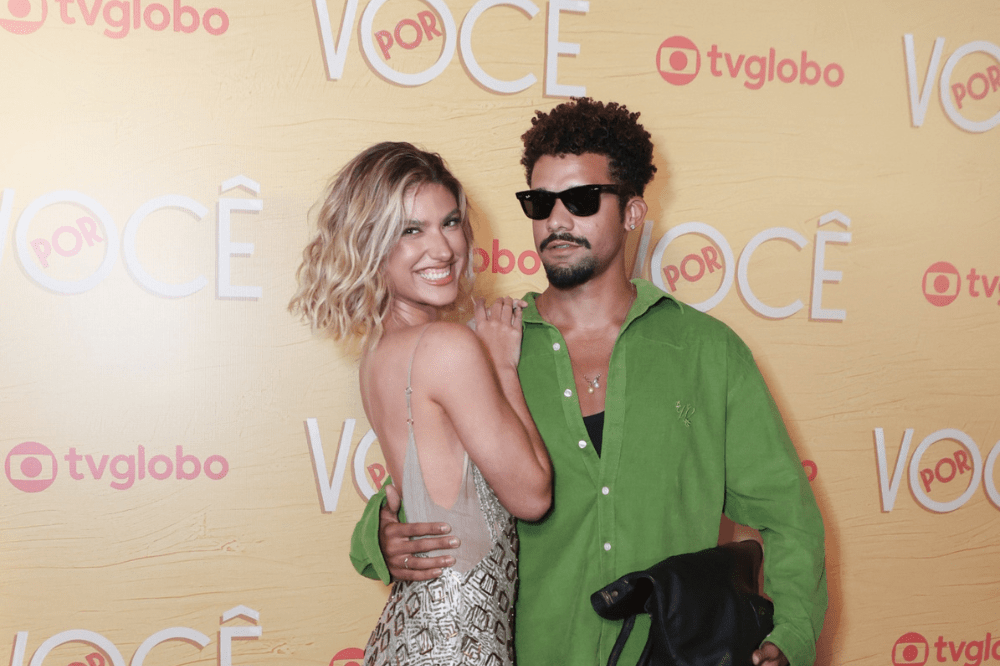 Um casal sorridente posa em frente a um painel amarelo com o logo tvglobo e VOCÊ POR. A mulher, loira de cabelo ondulado, usa um vestido prateado brilhante. O homem, de barba e cabelo crespo, veste camisa verde e óculos escuros, abraçando a mulher pela cintura