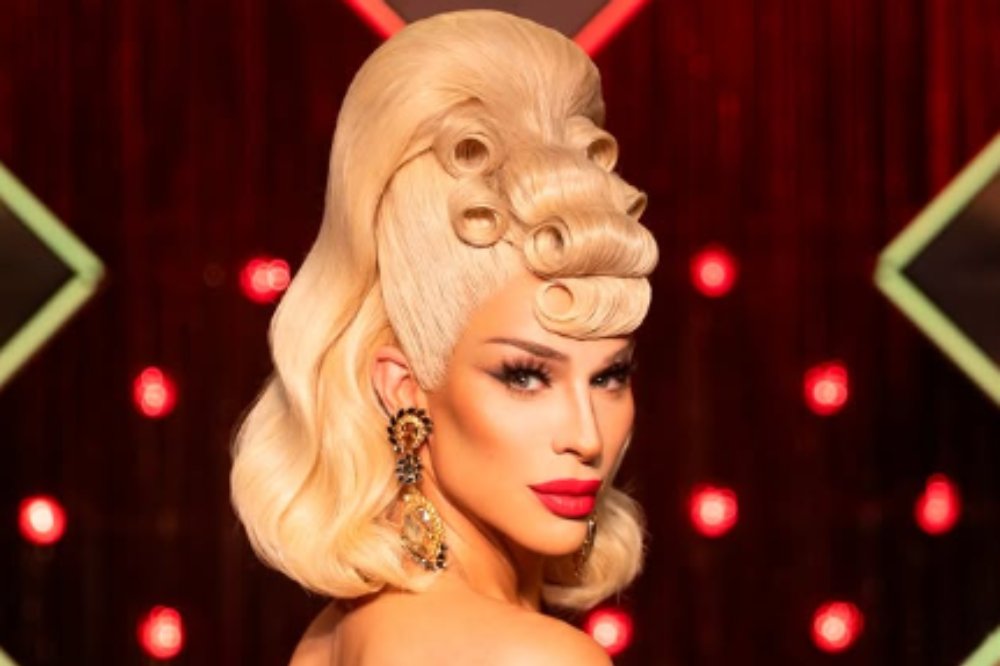 Drag queen loira com maquiagem marcante, batom vermelho e brincos grandes, olhando para a câmera. Seu cabelo tem um topete alto com cachos enrolados na frente. O fundo é escuro com luzes vermelhas desfocadas e formas geométricas verdes