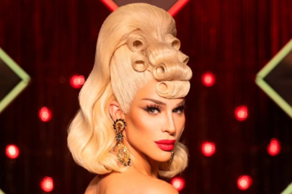 Drag queen loira com maquiagem marcante, batom vermelho e brincos grandes, olhando para a câmera. Seu cabelo tem um topete alto com cachos enrolados na frente. O fundo é escuro com luzes vermelhas desfocadas e formas geométricas verdes