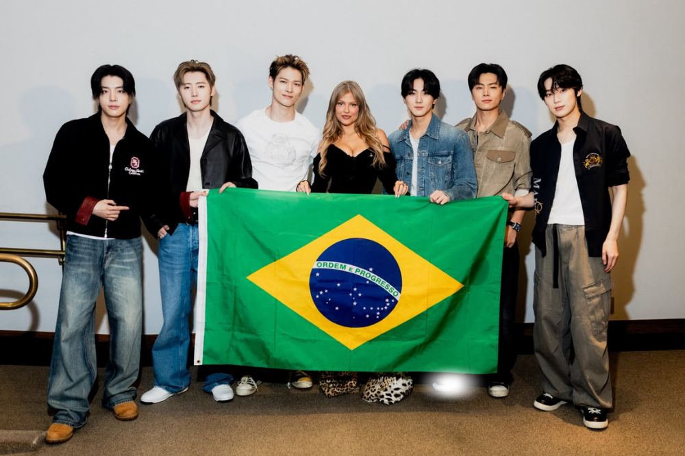 Sete pessoas, seis homens e uma mulher, posam sorrindo segurando uma bandeira do Brasil. Os homens vestem jaquetas e calças jeans ou cargo, e a mulher usa um top preto com estampa de onça na calça. Eles estão em um ambiente interno com parede clara e piso de carpete marrom