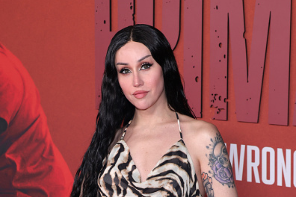 Mulher de cabelos pretos longos e lisos, maquiagem marcante, usando um vestido frente única com estampa de zebra, posa sorrindo levemente. Ela exibe tatuagens no braço esquerdo, em frente a um fundo vermelho com letras brancas desfocadas