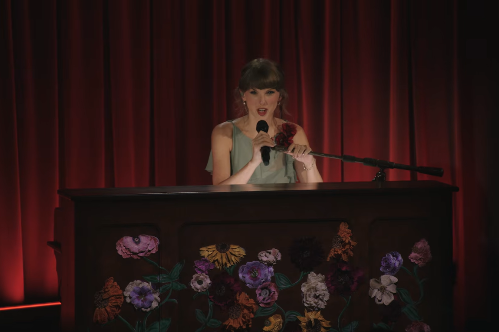 Taylor Swift, com um vestido azul claro, canta em um microfone enquanto segura uma rosa vermelha, atrás de um piano escuro decorado com flores coloridas, em um palco com cortinas vermelhas