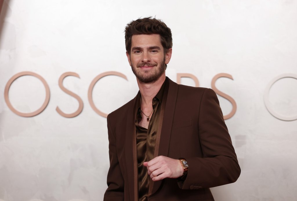 Andrew Garfield sorrindo, com barba e cabelo castanho, vestindo um terno marrom e camisa de seda, com um colar e relógio. Ao fundo, letras grandes e claras formam OSCARS