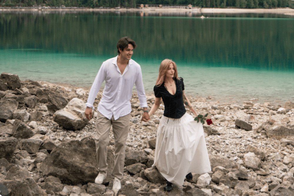 Casal sorridente, homem de camisa branca e calça cáqui, mulher de blusa preta e saia longa branca segurando uma rosa vermelha, caminhando de mãos dadas sobre pedras na margem de um lago de águas claras e esverdeadas, com floresta ao fundo