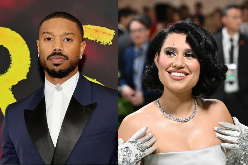 Michael B. Jordan, com barba e bigode, usa terno azul-marinho e broche prateado. Ao lado, uma mulher sorridente com cabelo escuro ondulado, colar de diamantes e luvas brancas bordadas