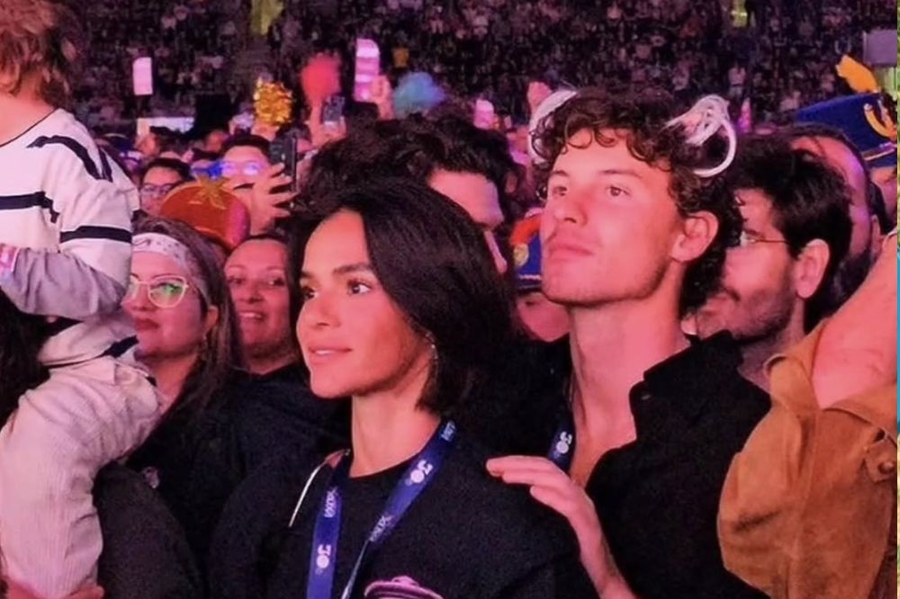 Bruna Marquezine, com cabelo escuro e liso, usa camiseta preta e crachá azul, ao lado de Shawn Mendes, de cabelo cacheado e camisa preta, com orelhas de gato brancas na cabeça. Ambos olham para cima em meio a uma multidão em um show, com luzes coloridas ao fundo