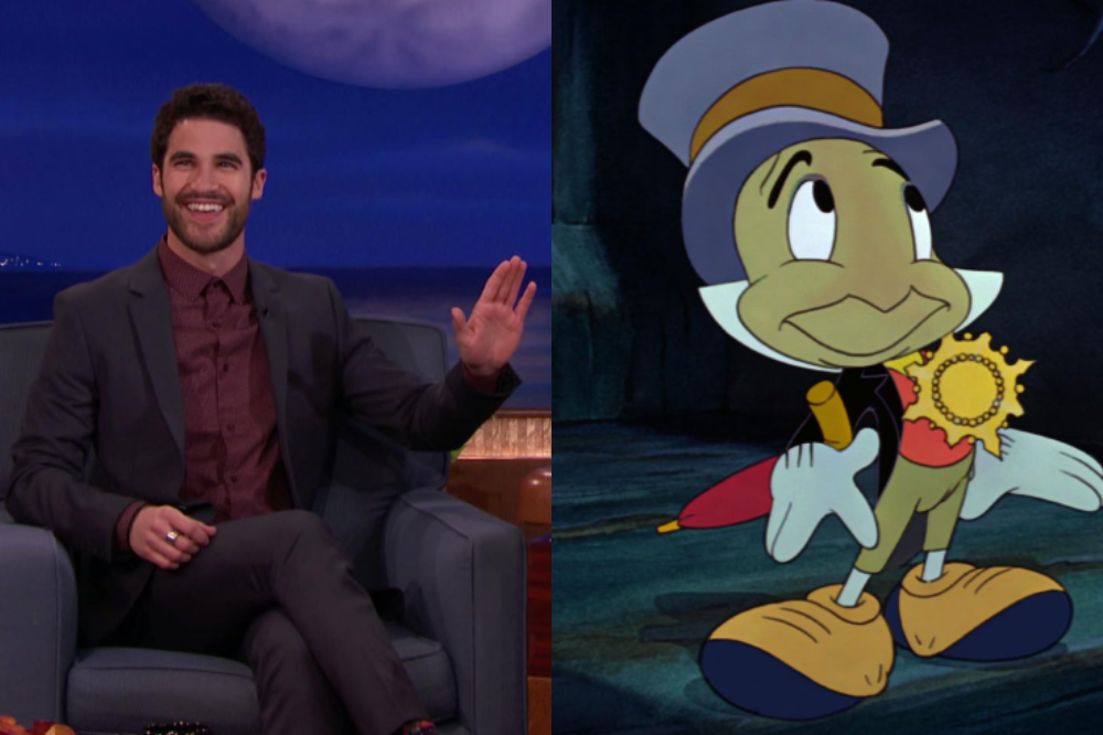 O ator Darren Criss sorri e acena, sentado em um sofá, ao lado do Grilo Falante, personagem de desenho animado, com chapéu e guarda-chuva