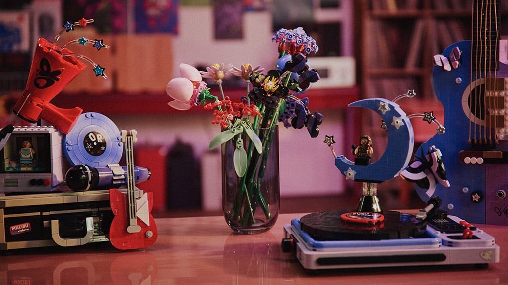 Cena com objetos de Lego: um megafone vermelho, uma televisão com boneca, uma guitarra vermelha, um vaso com flores coloridas, um toca-discos com um boneco em uma lua azul e um violão azul