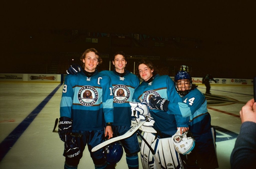 Quatro jovens jogadores de hóquei, três sorrindo, posam na pista de gelo. Usam uniformes azuis e turquesa com o logo South Dorothy Grizzlies e um urso pardo. Um deles é goleiro, segurando o taco e o capacete.