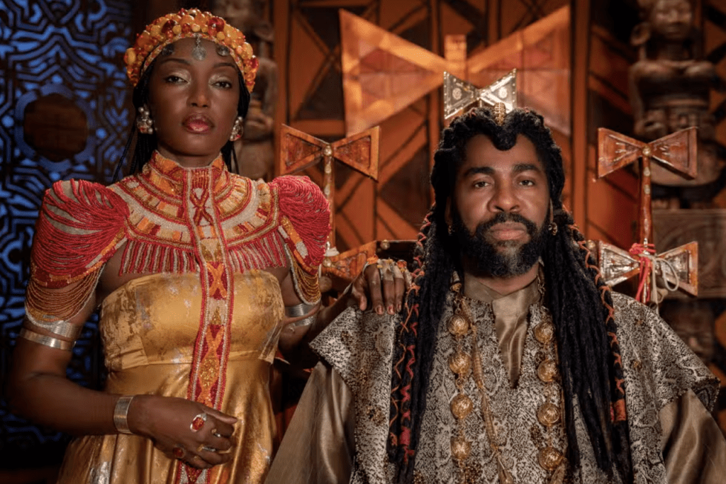 Uma mulher e um homem negros, vestidos com trajes cerimoniais africanos, posam com expressões sérias. Ela usa um vestido dourado com ombros e gola adornados por miçangas vermelhas e douradas, e uma coroa de miçangas. Ele tem dreadlocks longos, barba e veste uma túnica estampada com colares dourados. Ao fundo, elementos decorativos em madeira escura e dourada.