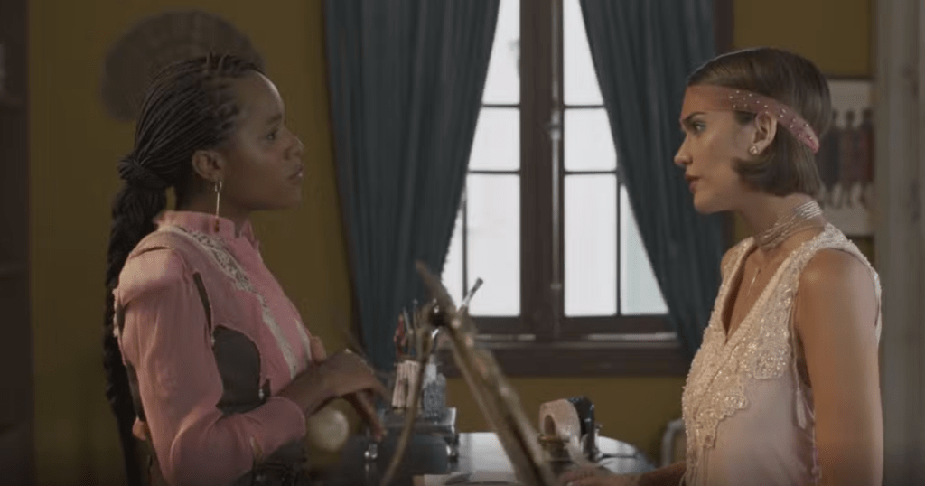 Duas mulheres conversam em um ambiente interno. À esquerda, uma mulher negra com tranças e blusa rosa com detalhes brancos. À direita, uma mulher branca de cabelo curto, faixa rosa na cabeça e vestido branco brilhante. Ambas se olham, com expressões sérias. Ao fundo, uma janela com cortinas azuis e uma parede amarela
