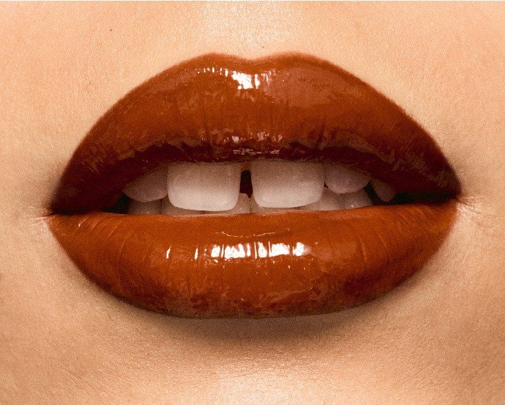 Boca de uma pessoa com lábios carnudos e brilhantes, maquiados com batom marrom avermelhado. Os dentes brancos da frente têm um pequeno espaço entre eles