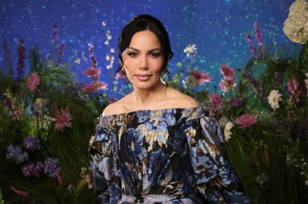 Mulher de pele clara, cabelo escuro preso com mechas brancas na frente, maquiagem leve, usando um vestido azul e dourado com ombros à mostra, posa sorrindo em um cenário de jardim com flores coloridas e fundo azul estrelado