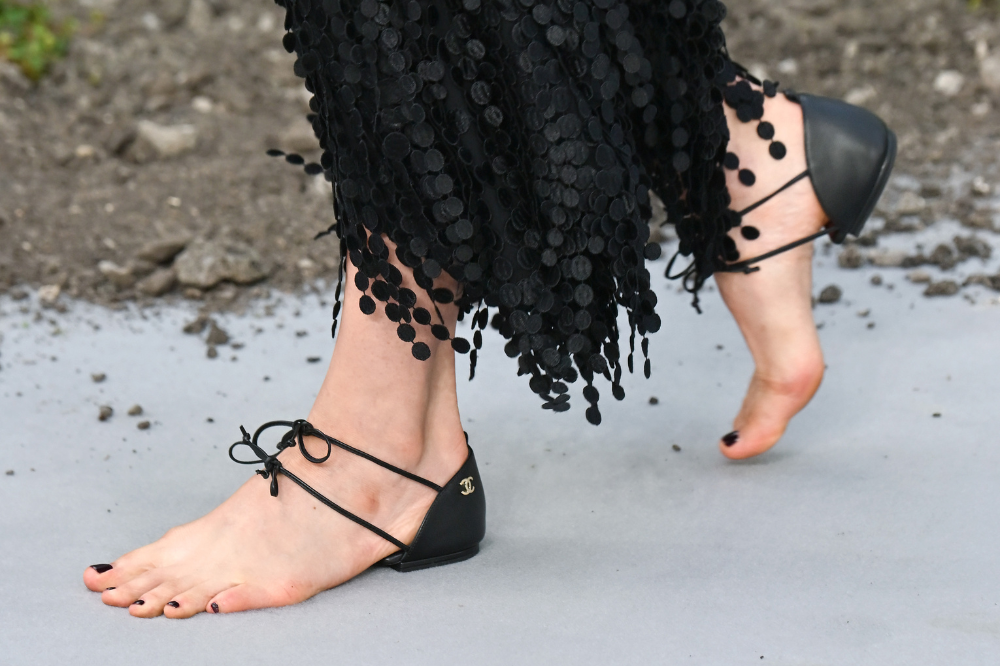 Pés de uma pessoa com esmalte preto e sandálias rasteiras pretas com tiras amarradas nos tornozelos, sobre um piso cinza com terra e pedras. A pessoa veste uma saia preta com detalhes circulares pendurados
