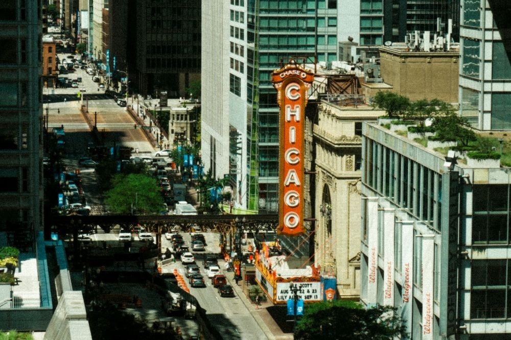 Vista aérea da rua State Street em Chicago, com o letreiro vertical vermelho do Teatro Chicago em destaque. Carros e ônibus trafegam pela rua, ladeada por edifícios altos e modernos. Um viaduto de trem elevado atravessa a imagem, e faixas da Walgreens são visíveis em um prédio à direita