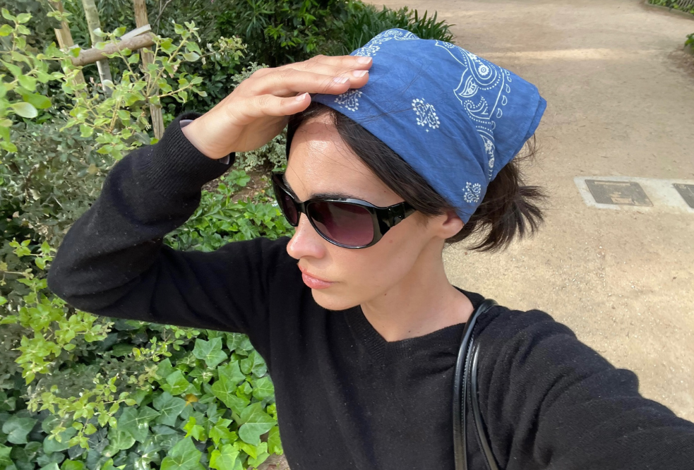 Mulher de óculos escuros e bandana azul com estampa branca, usando blusa preta, protegendo os olhos do sol com a mão, em um caminho de terra com vegetação verde ao lado