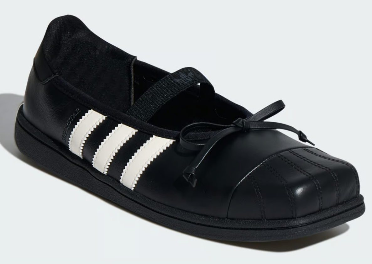 Sapatilha preta Adidas Superstar, com três listras brancas serrilhadas na lateral, bico quadrado com costuras verticais e laço decorativo na frente. Uma tira elástica preta com o logo Adidas atravessa o peito do pé