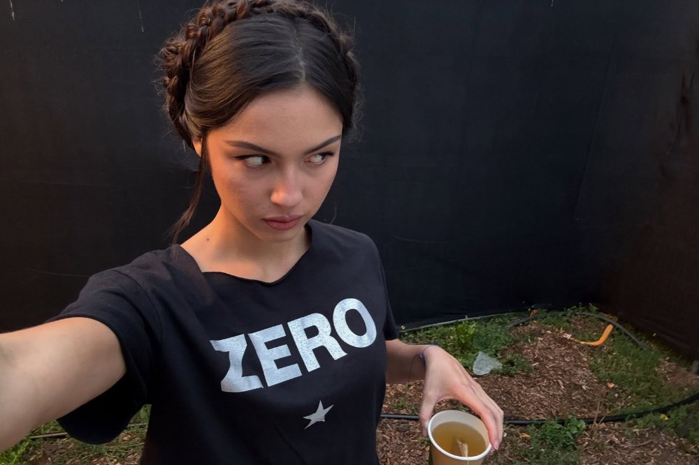 Jovem mulher com tranças no cabelo, vestindo camiseta preta com a palavra ZERO e uma estrela branca, segurando um copo com chá, olhando para o lado com expressão séria. O fundo é uma parede preta e chão de terra com grama