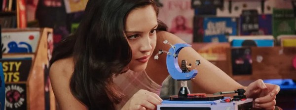 Jovem mulher de cabelos escuros e ondulados, vestindo regata clara, observa atentamente um toca-discos de brinquedo com um enfeite de lua azul. Ela está em um quarto com paredes cobertas por pôsteres e prateleiras cheias de discos de vinil