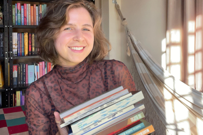 Mulher jovem de cabelo castanho curto e blusa de oncinha marrom sorri, segurando uma pilha de livros. Ao fundo, uma estante cheia de livros e uma rede listrada com luz solar entrando pela janela