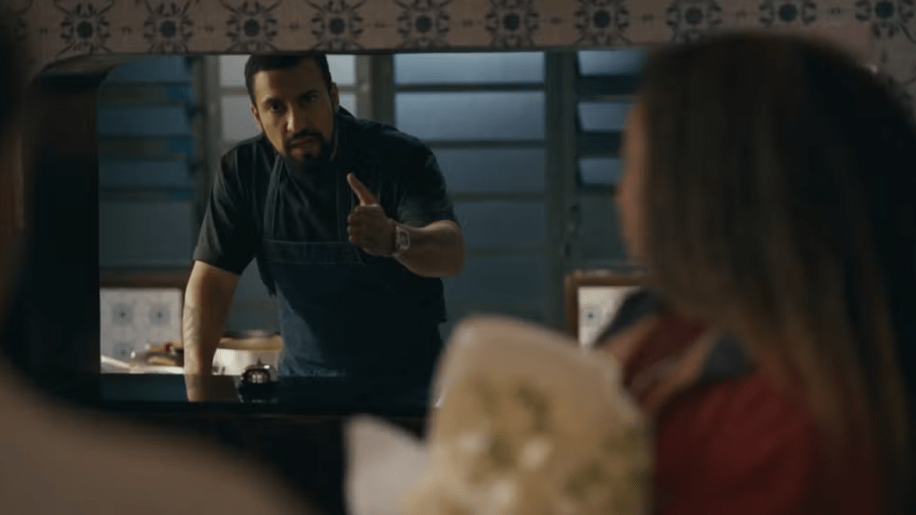 Um homem barbudo, de camisa escura e avental, faz um sinal de joinha com a mão direita, olhando para uma mulher de costas, com cabelos longos, em um ambiente interno com azulejos decorados