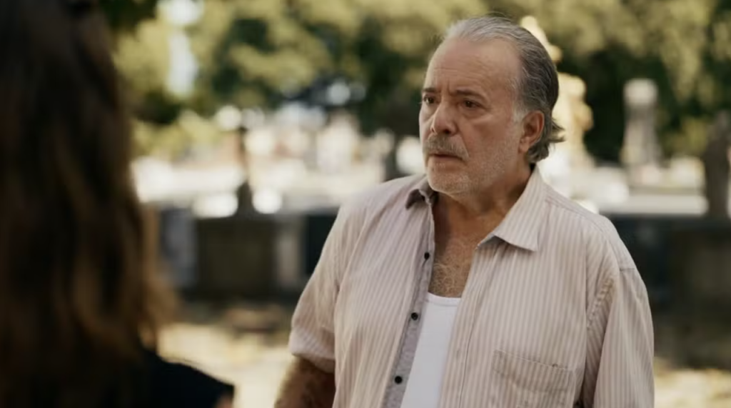 Homem idoso de cabelos grisalhos e barba rala, vestindo camisa listrada clara e camiseta branca, olha para a esquerda com expressão séria. Ao fundo, árvores e lápides de um cemitério, com uma pessoa de costas à esquerda