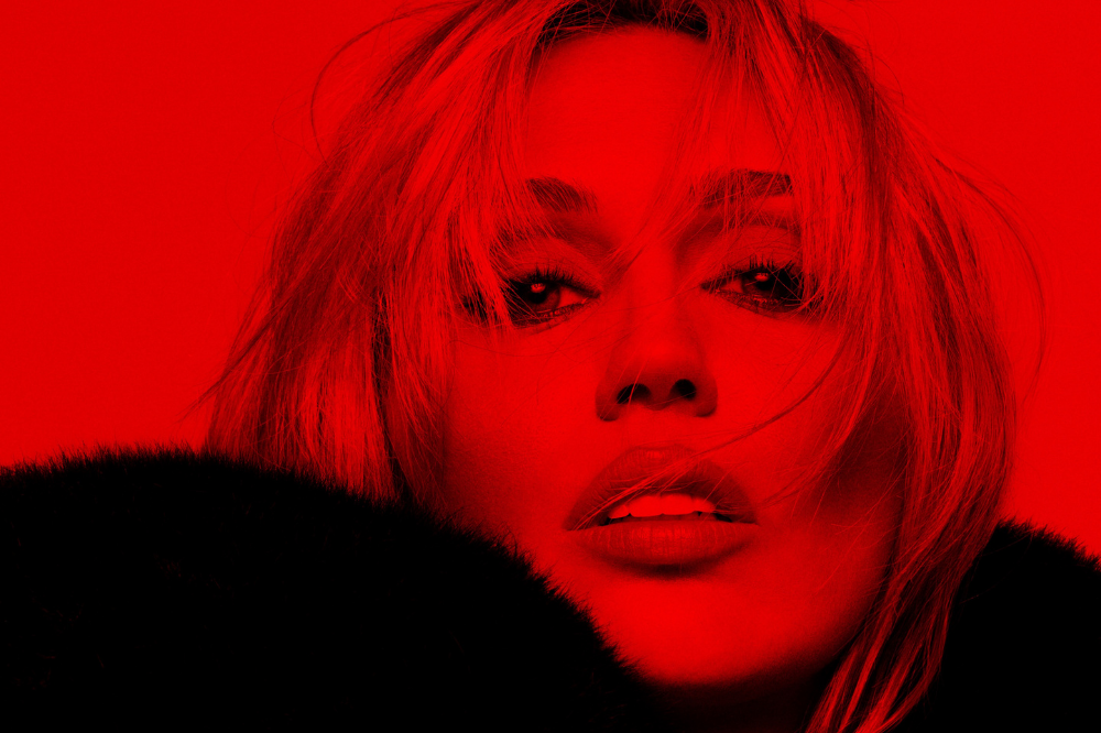 Rosto de uma mulher com cabelo loiro bagunçado, maquiagem escura nos olhos e lábios entreabertos, em um fundo vermelho vibrante. Ela usa uma gola de pele escura que cobre parte do ombro esquerdo. A imagem tem um filtro vermelho intenso, criando um efeito dramático