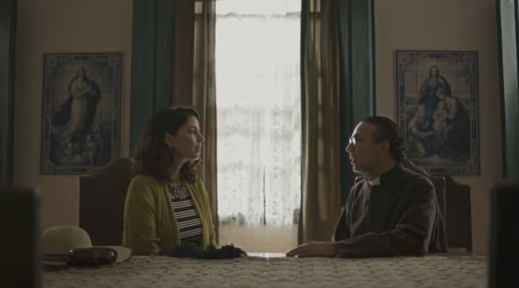 Uma mulher de blusa listrada e casaco claro conversa com um padre de batina preta, sentados à mesa em um ambiente interno com quadros religiosos na parede e uma janela ao fundo