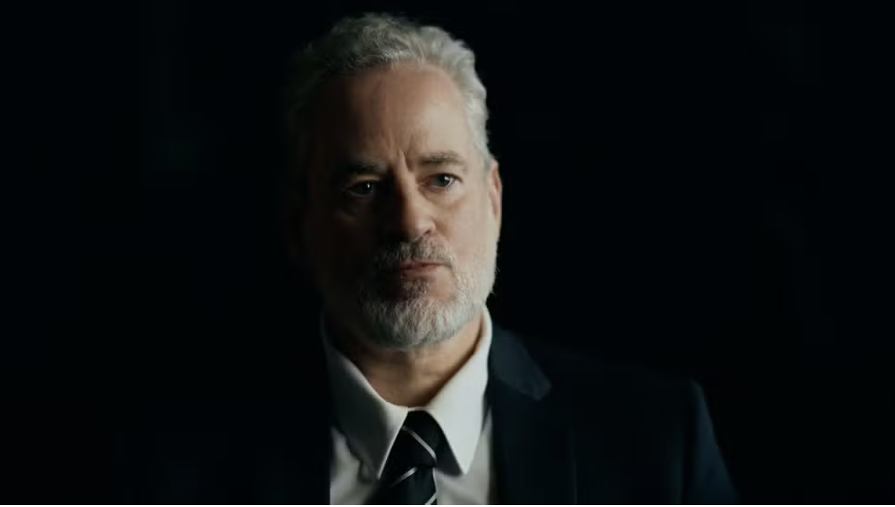 Homem de meia-idade com cabelo e barba grisalhos, vestindo terno escuro e gravata listrada, olhando seriamente para frente, com iluminação dramática no rosto e fundo escuro