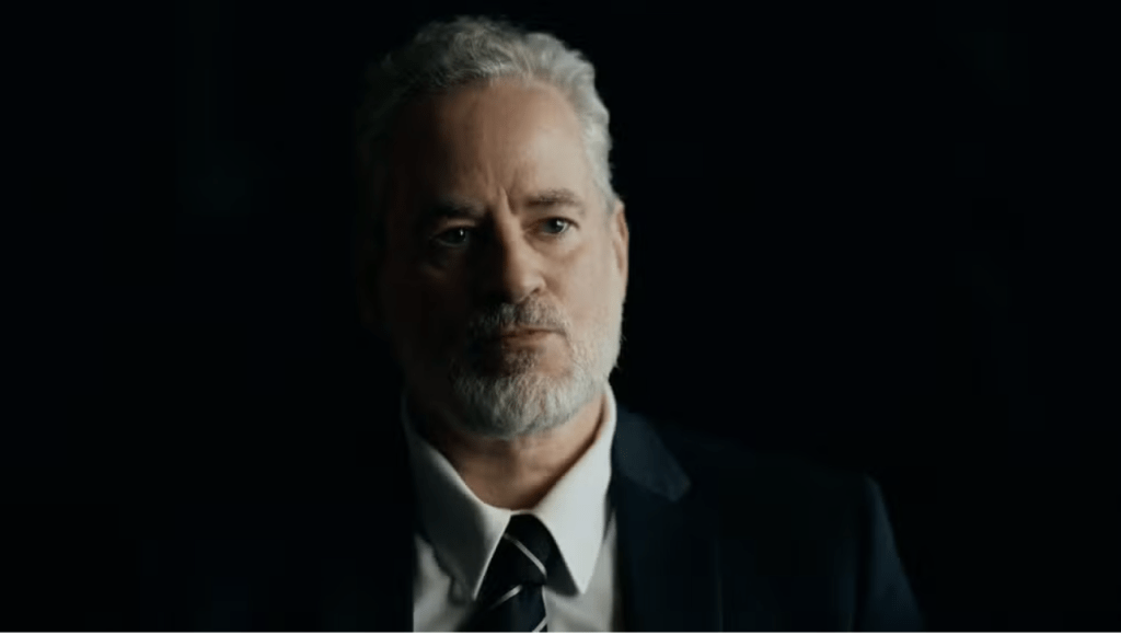 Homem de meia-idade com cabelo e barba grisalhos, vestindo terno escuro e gravata listrada, olhando seriamente para frente, com iluminação dramática no rosto e fundo escuro